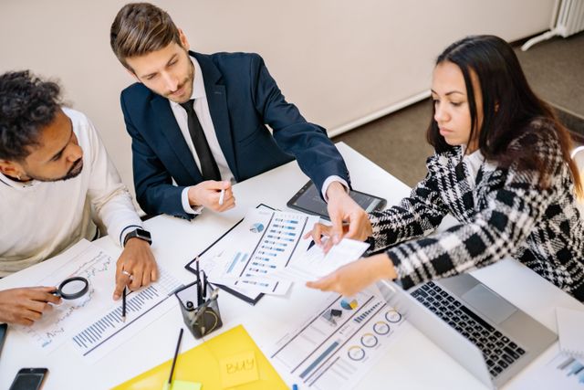 Business-First - Nwaresoft Private Limited - Nwaresoft Private Limited Three professionals at a white table review charts and reports; one points with a pen to a graph.“