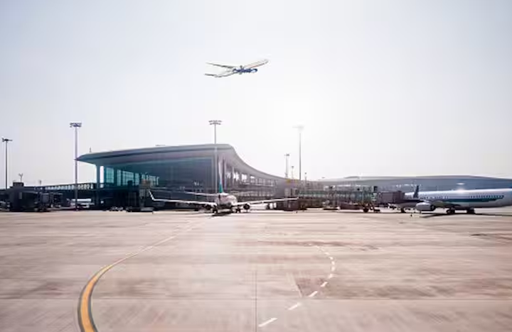 - - Airport terminal and parked airplanes on the tarmac, with a small plane taking off in the bright sky above.