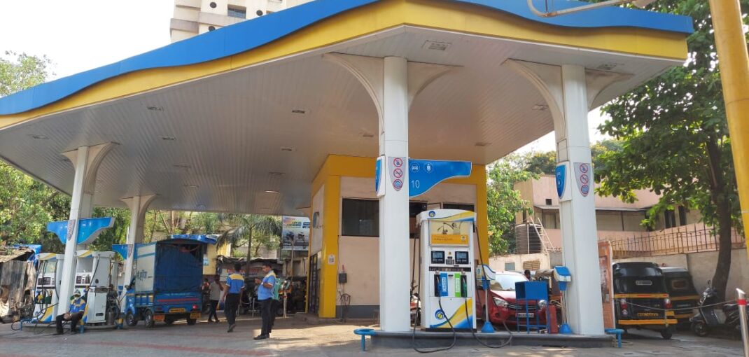 - - Petrol station with a blue-and-yellow canopy, fuel pumps, and attendants; auto rickshaws and a tall building visible in the background.