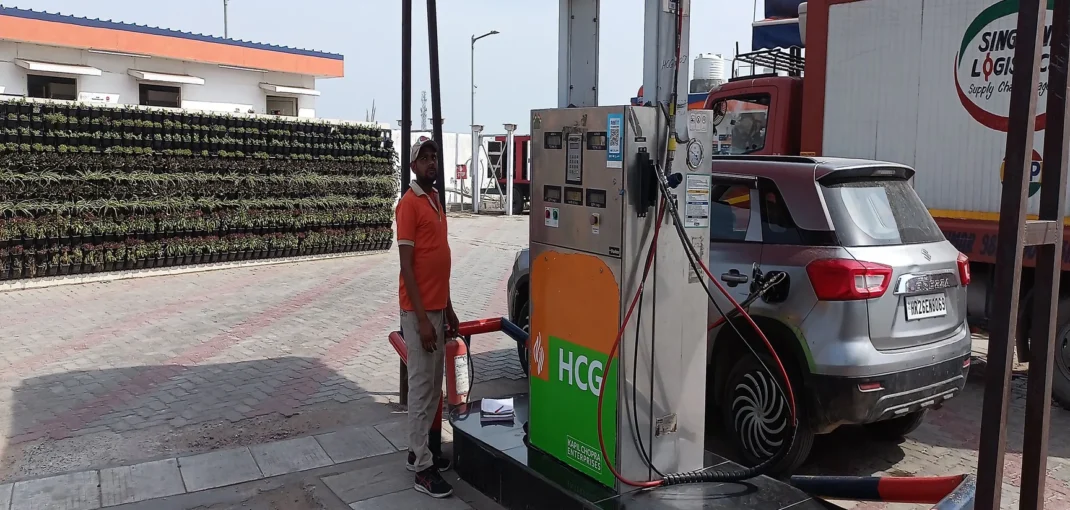 - - A man in an orange shirt stands beside a fuel pump connected to a silver SUV at an outdoor station, with a busin area and plant wall in the background.