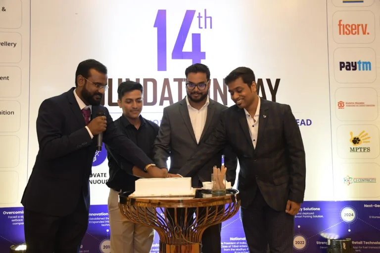 - Four men in suits cut a cake on a stage during a foundation day celebration with a large '14th Foundation Day' backdrop behind them.